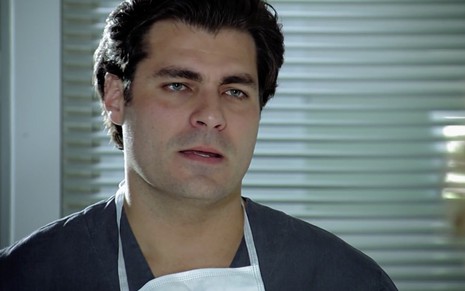O ator Thiago Lacerda como Lúcio com um capote azul e uma máscara cirúrgica branca no queixo, ele está em um ambiente branco de um hospital em cena de A Vida da Gente