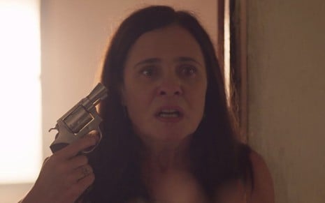 A atriz Adriana Esteves segura uma arma como a vilã Thelma de Amor de Mãe e faz cara de maluca em cena