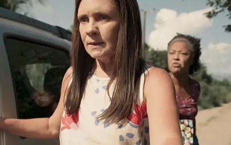 A atriz Adriana Esteves como Thelma em primeiro plano, ela segura a porta de um carro com expressão de raiva, enquanto Mariana Nunes como Rita grita ao fundo da cena de Amor de Mãe