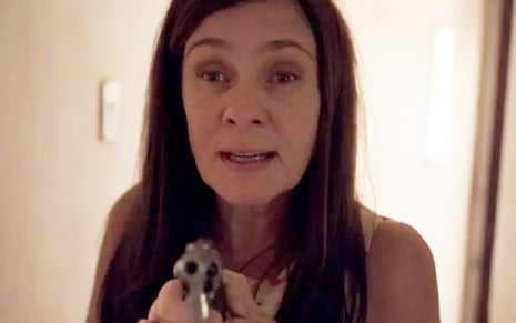 A atriz Adriana Esteves caracterizada como Thelma, ela tem expressão de descontrole e segura uma arma com as duas mãos em cena de Amor de Mãe