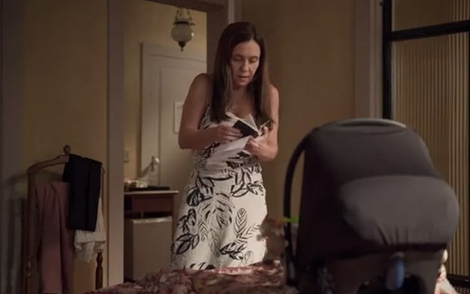 A atriz Adriana Esteves caracterizada como Thelma mexe em um passaporte ao lado de um carrinho de bebê em cena de Amor de Mãe