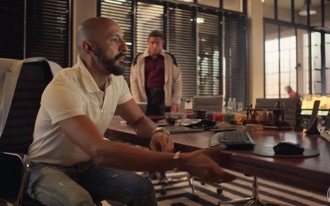 O ator Irandhir Santos olha para computador em cena de Amor de Mãe em que Tuca Andrada aparece desfocado