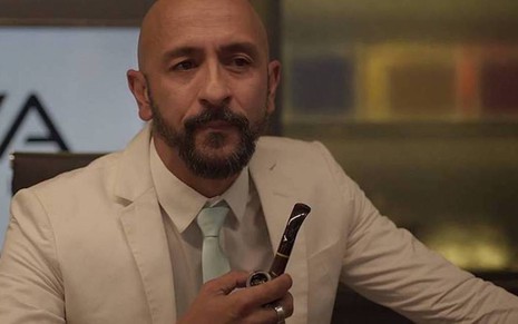 O ator Irandhir Santos, vestido com um terno claro, segura um cachimbo em cena da novela Amor de Mãe, da TV Globo