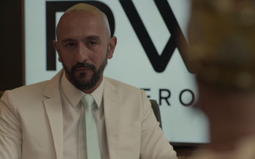 O ator Irandhir Santos com um terno claro conversa com Jéssica Ellen no cenário do escritório de seu personagem na novela das nove