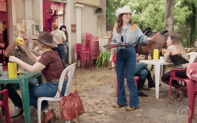 A atriz Arieta Corrêa está com uma roupa de cowboy, segurando uma bandeja redonda em um bar com várias mesas e até mesmo um cavalo ao fundo, ela está caracterizada como Leila em cena de Amor de Mãe
