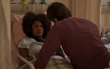Jéssica Ellen deitada em leito de hospital conversa com Chay Suede (de costas) em cena da novela das nove da TV Globo