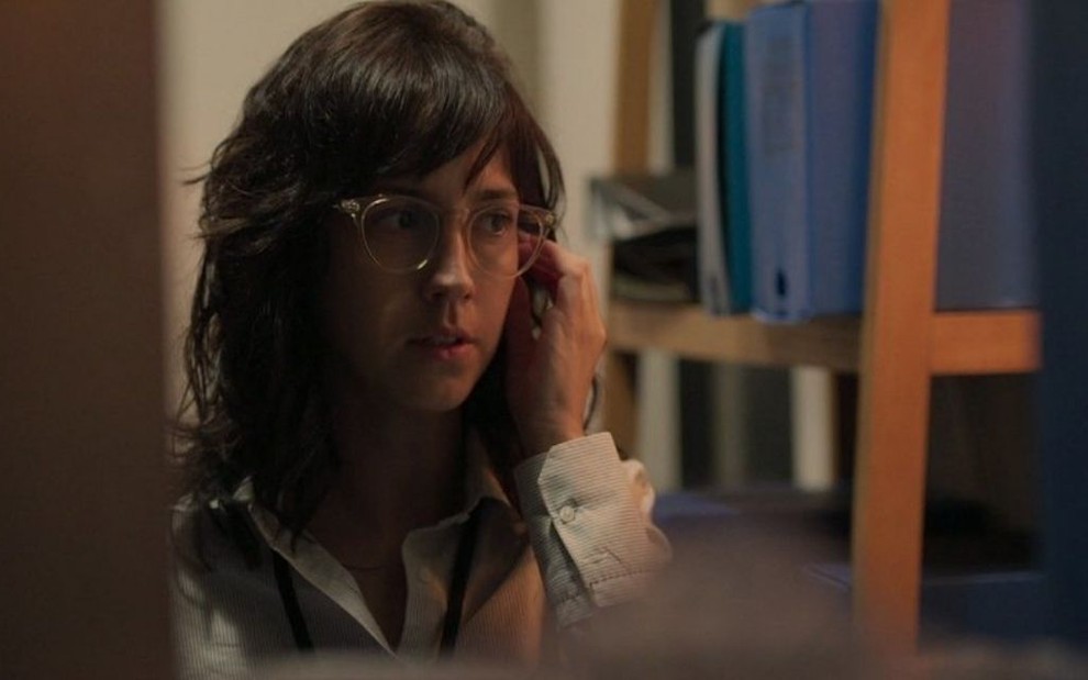 A atriz Camila Márdila, com um óculos de armação transparente, está escondida atrás de uma estante enquanto fala no celular em cena de Amor de Mãe