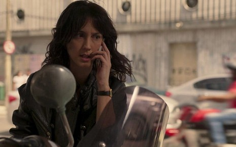 Camila Márdila segura um celular ao lado da orelha esquerda, enquanto está sentada em uma moto, caracterizada como a Amanda em cena de Amor de Mãe