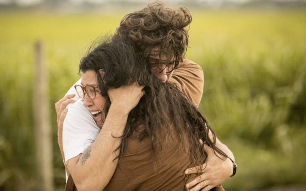 O ator Chay Suede como Danilo abraça Regina Casé como Lurdes em uma estrada de terra com um matagal ao fundo em cena de Amor de Mãe