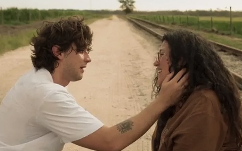 O ator Chay Suede como Danilo à esquerda leva as mãos ao rosto de Regina Casé como Lurdes à direita  em frente a uma estrada de terra em cena de Amor de Mãe