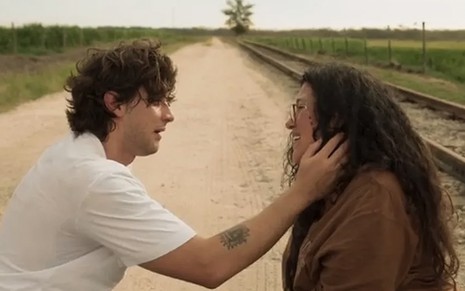 O ator Chay Suede à esquerda como Danilo segura o rosto de Regina Casé, a Lurdes, com carinho em frente a uma estrada de terra em cena de Amor de Mãe