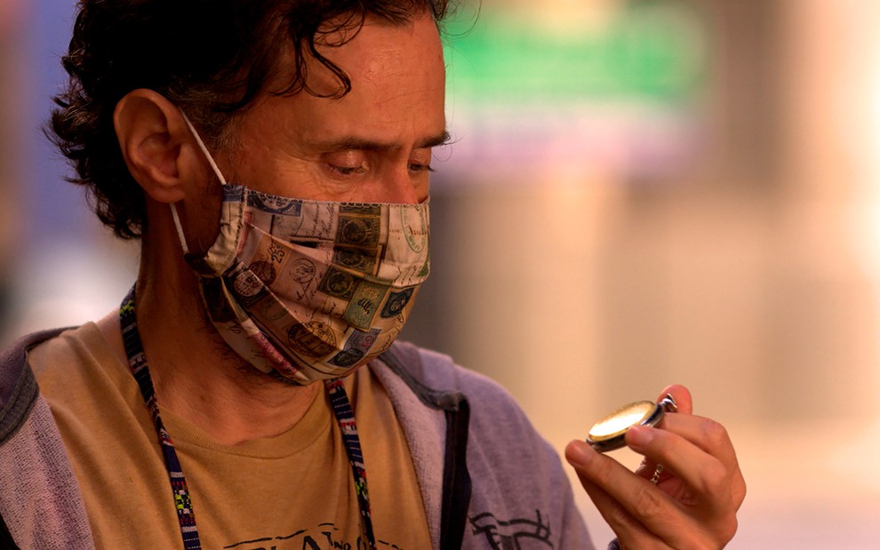 O ator Enrique Dias com uma máscara colorida segura um objeto redondo com a mão direita caracterizado como Durval em cena de Amor de Mãe