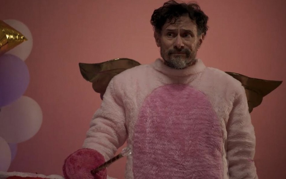 Em frente a um fundo rosa, o ator Enrique Diaz está fantasiado de unicórnio, mas com a cabeça livre, em cena de Amor de Mãe