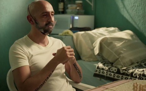 O ator Irandhir Santos, sentado, usando uma máscara transparente, em cena como Álvaro em Amor de Mãe