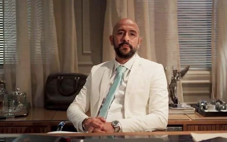 O ator Irandhir Santos, sentado, com um terno branco e gravata azul, em cena como Álvaro em Amor de Mãe