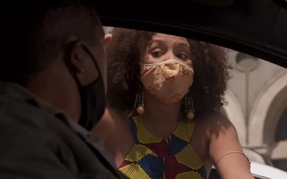 A atriz Jéssica Ellen invade um carro pela janela com uma máscara no rosto e uma roupa florida, há um homem dentro do carro, na penumbra, em cena de Amor de Mãe