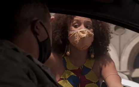 A atriz Jéssica Ellen invade um carro pela janela com uma máscara no rosto e uma roupa florida, há um homem dentro do carro, na penumbra, em cena de Amor de Mãe