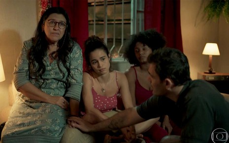 Regina Casé em cena de Amor de Mãe: caracterizada como Lurdes, personagem está sentada no braço do sofá; do seu lado está Nanda Costa caracterizada como Érica e Jéssica Ellen caracterizada como Camilla