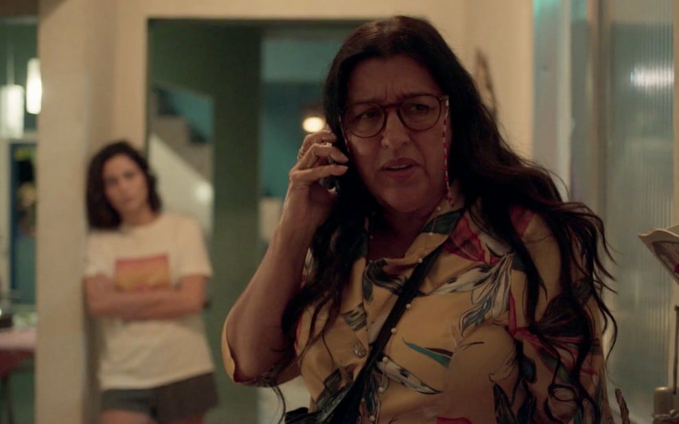 Regina Casé em cena de Amor de Mãe: personagem usa o celular e faz cara de confusão e Nanda Costa aparece ao fundo