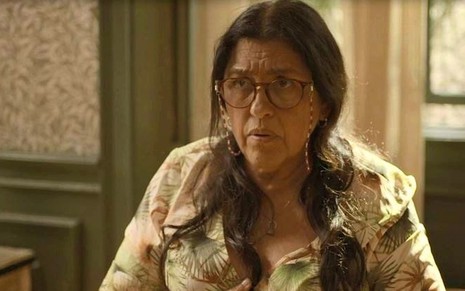 Regina Casé usa vestido florido e óculos em cena de Amor de Mãe na qual faz uma expressão de estranhamento