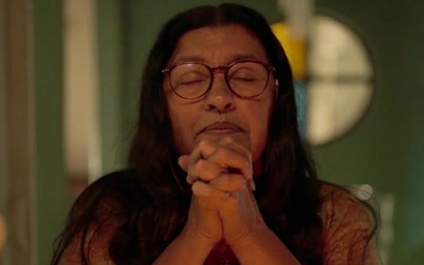Regina Casé em cena de Amor de Mãe: caracterizada como Lurdes, personagem está de olhos fechados e ora