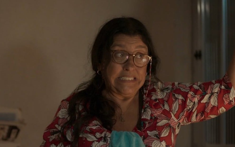 A atriz Regina Casé usa óculos, vestido florido e faz uma expressão engraçada em cena da novela Amor de Mãe, da Globo