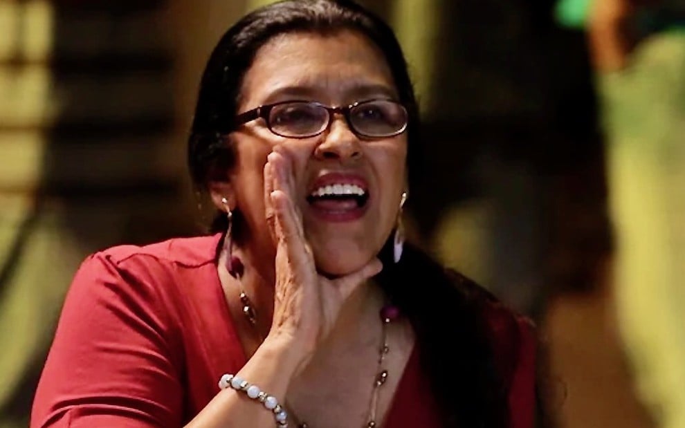 A atriz Regina Casé usa uma blusa vermelha e coloca a mão na altura da boca para dar um aviso em cena da novela Amor de Mãe