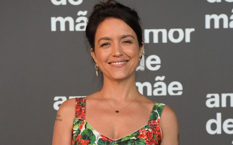 A escritora Manuela Dias com um vestido florido de alças e com decote no colo em frente a um backdrop em que se lê Amor de Mãe durante a festa de lançamento da novela