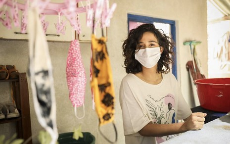A atriz Nanda Costa como Érica usa uma máscara branca enquanto posa em uma área de serviço com várias máscaras em um varal em cena de Amor de Mãe