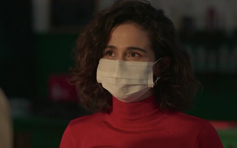A atriz Nanda Costa usa uma máscara branca e uma blusa de gola alta vermelha caracterizada como Érica em cena de Amor de Mãe
