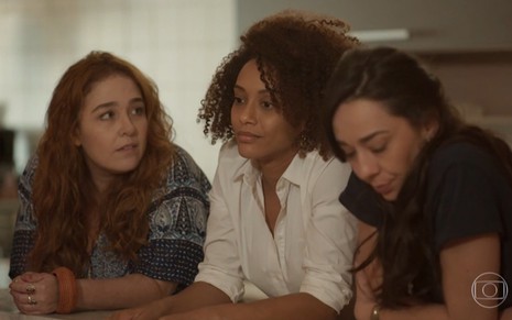 Debora Lamm, Taís Araujo e Clarissa Kiste gravam lado a lado apoiadas em mesa em cena da novela Amor de Mãe