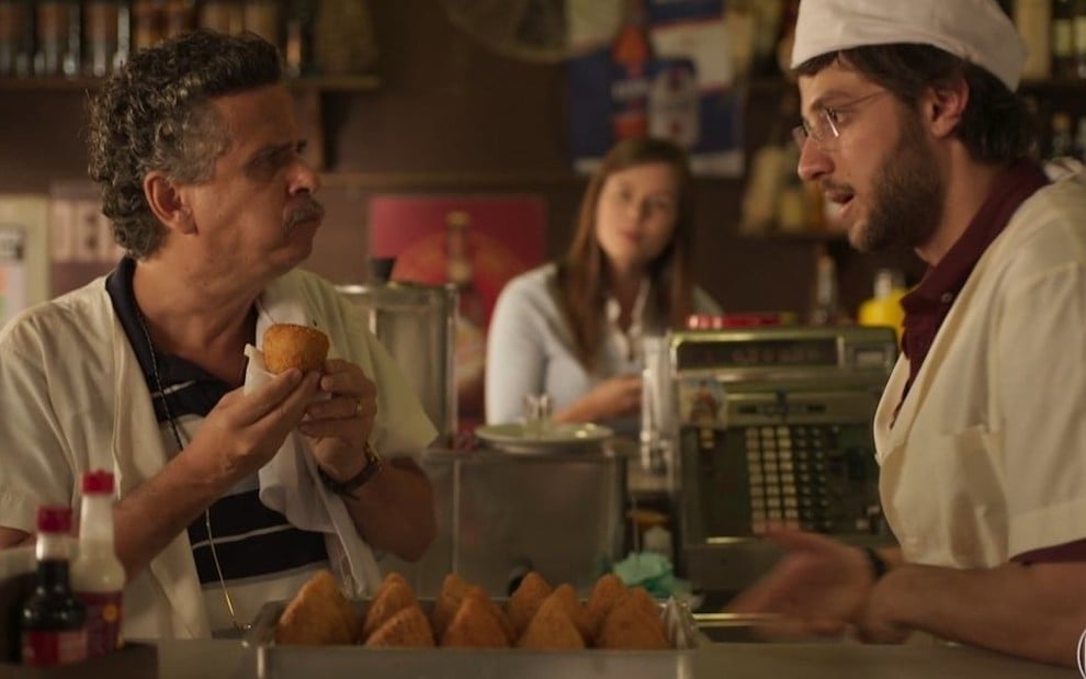 O ator Rodolfo Vaz come uma coxinha na frente de Chay Suede no cenário do bar de seu personagem em Amor de Mãe, novela das nove da Globo