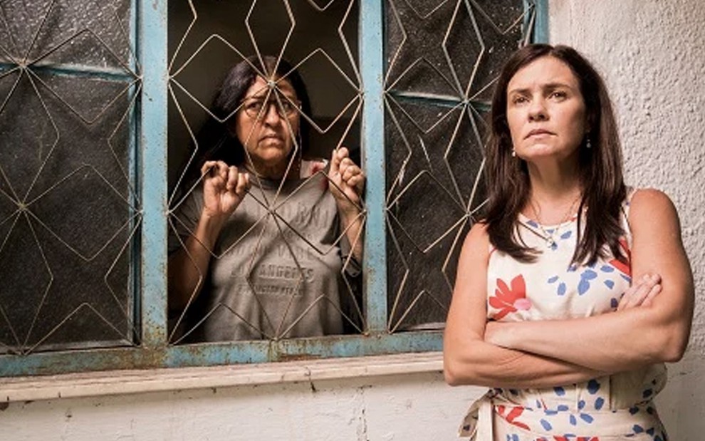 Atrás das grades de uma janela, Regina Casé como Lurdes observa Adriana Esteves, a Thelma, que está de braços cruzados em primeiro plano na cena de Amor de Mãe