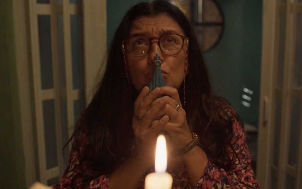 A personagem Lurdes (Regina Casé) reza para santa e em frente a vela acesa em cena da novela Amor de Mãe, da Globo