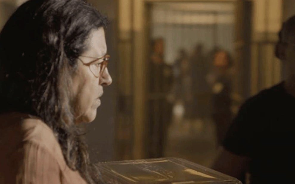 A atriz Regina Casé, de perfil, segura uma embalagem com um bolo de chocolate caracterizada como a personagem Lurdes em Amor de Mãe, ela está em uma fila em uma prisão