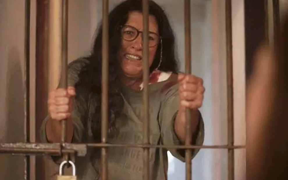 A atriz Regina Casé como Lurdes está presa em uma jaula, ela segura às barras de ferro e tem expressão de desespero em cena de Amor de Mãe