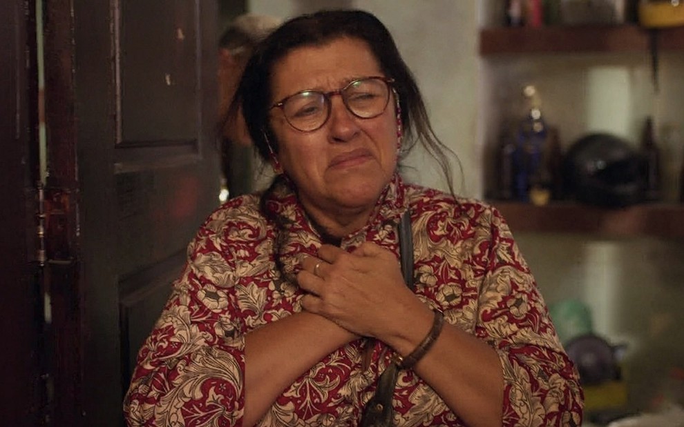 A atriz Regina Casé chora com as mãos entrelaçadas no peito, ela está caracterizada como Lurdes em cena de Amor de Mãe