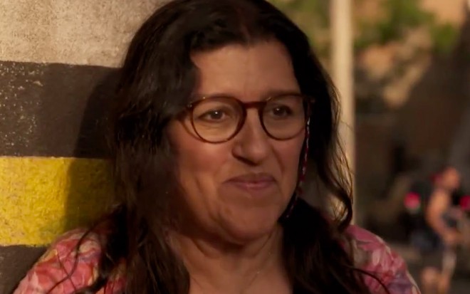 A atriz Regina Casé, com óculos e uma roupa rosa, sorri timidamente caracterizada como a Lurdes em cena de Amor de Mãe