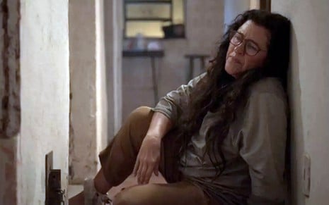 A atriz Regina Casé sentada no chão, com uma perna acorrentada a um grilhão, como dona Lurdes em cena de Amor de Mãe