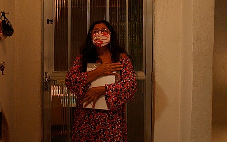 A atriz Regina Casé com uma máscara vermelha estampada abraça uma pasta cheia de documentos como a Lurdes em cena de Amor de Mãe