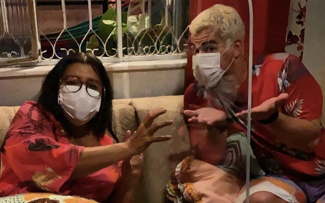 A atriz Regina Casé à esquerda como Lurdes separada por um acrílico no sofá de Thiago Martins à direita como Ryan durante as gravações de Amor de Mãe