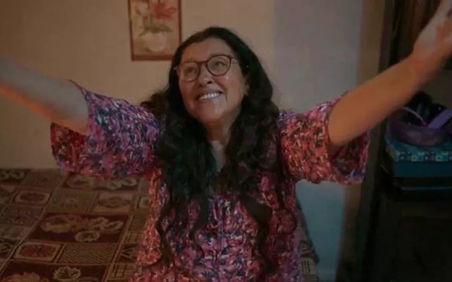A atriz Regina Casé, com os braços levantados para o céu, em cena como Lurdes em Amor de Mãe