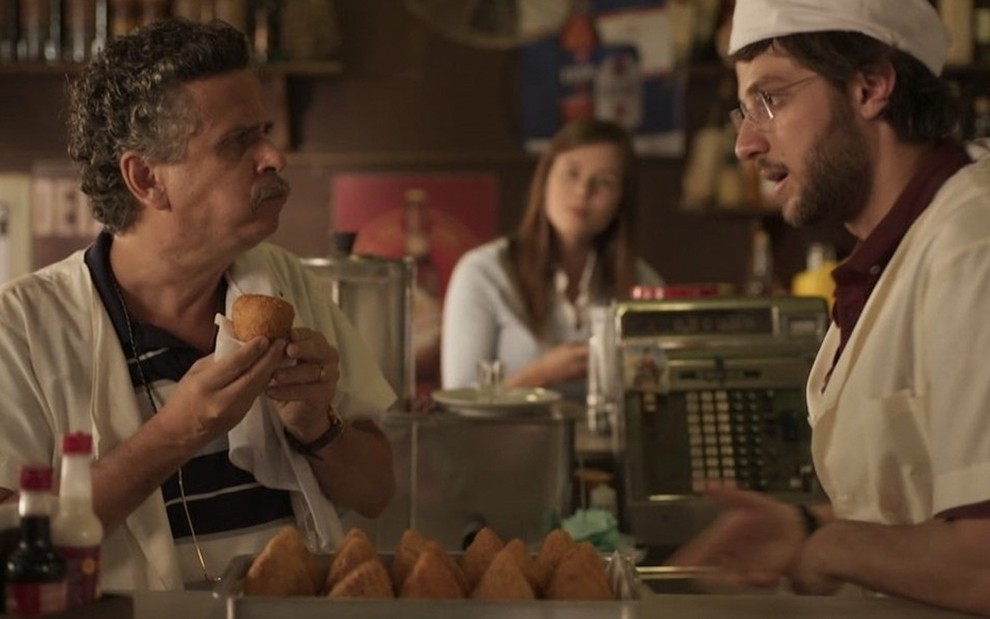 O ator Rodolfo Vaz à esquerda como Álvaro, ele está de boca cheia e segura uma coxinha com a parte da bunda virada para cima, ele olha para Chay Suede, o Danilo, que está de doma e chapéu de cozinheiro em cena de Amor de Mãe