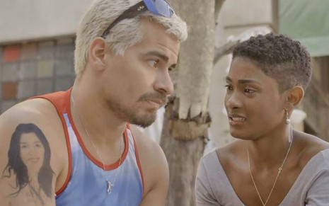 O ator Thiago Martins, que usa camiseta regata e óculos escuros na cabeça, conversa com Erika Januza, que faz cara de choro, em cena de Amor de Mãe