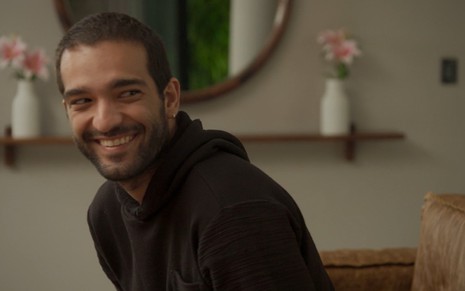 Humberto Carrão grava com casaco cinza escuro e sorriso no rosto em cena como Sandro de Amor de Mãe