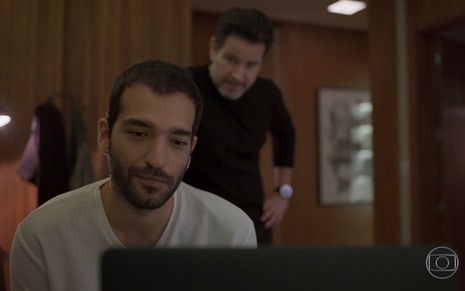 Humberto Carrão grava com expressão séria em frente ao computador, Murilo Benício está de braços cruzados atrás dele