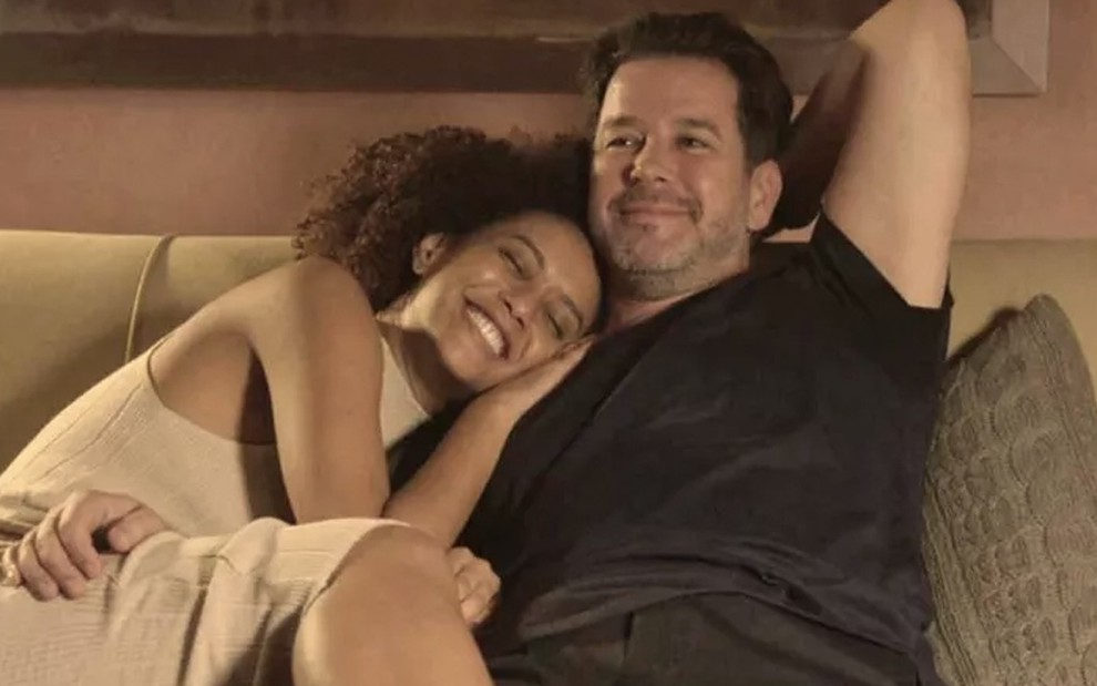 Taís Araujo, caracterizada como Vitória, deitada sobre o peito de Murilo Benício, o Raul, enquanto os dois estão sentados em um sofá, ambos com expressão de felicidade, em cena de Amor de Mãe