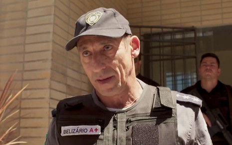 O ator Tuca Andrada fardado como um policial militar, com um boné da corporação, caracterizado como o Belisário de Amor de Màe
