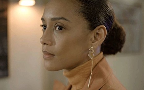 A atriz Taís Araujo exibe uma expressão forte em cena que está com cabelo preso e uma blusa de gola alta em Amor de Mãe
