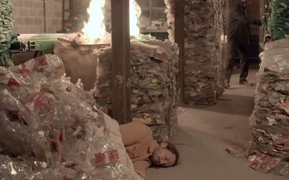 O ator Vladimir Brichta caracterizado como o Davi caído no chão, desacordado, em meio a pilhas de lixo que pegam fogo em cena de Amor de Mãe